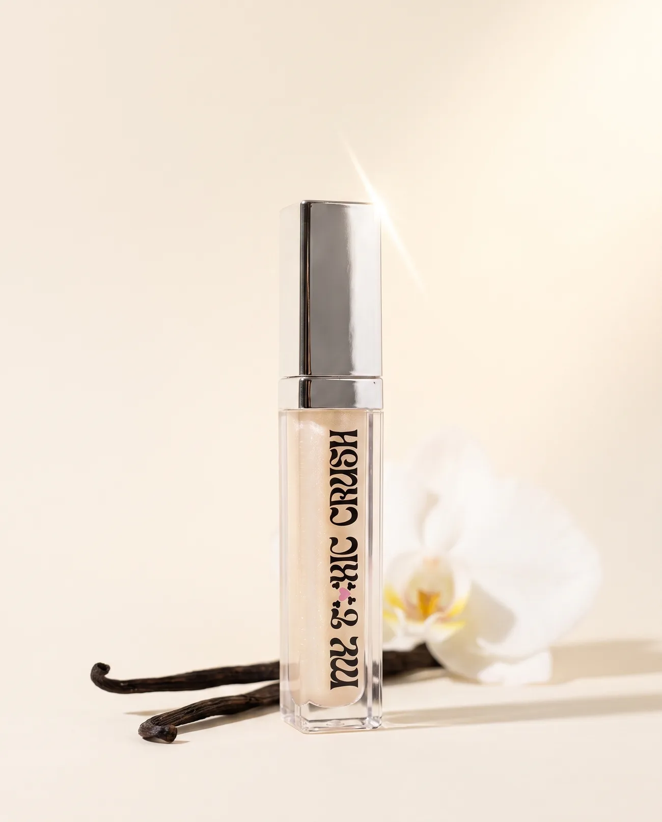 Vanilla lie lip gloss standing beside vanilla pods and a white orchid on a cream backdrop